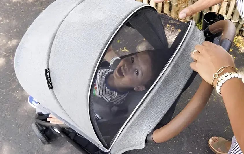 A smiling baby looking up through the mesh window of an Evenflo stroller canopy while a caregiver peeks inside.